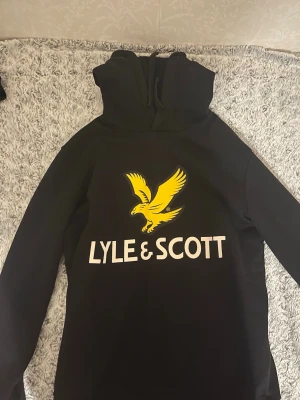 Svart hoodie priset kan diskuteras - Svart hoodie från Lyle & Scott med stor gul örn och vit logga på bröstet. Klassisk modell med huva och dragsko. Perfekt för dig som gillar streetwear och vill sticka ut med en tydlig logga. Mjuk och skön bomullsblandning.