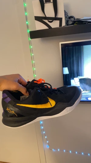 Kobe basketskor - Snygga Nike Kobe sneakers i svart med gula och lila detaljer samt vit sula. Skon har en låg profil och är tillverkad i textil och syntetmaterial. Klassisk Kobe-signatur på hälen och orange skosnören för extra pop. Perfekt för dig som gillar basketinspirerad stil. Skon har ett litet hål som ni ser på sista bilden men de påverkar inte något när man spelar med dem. Och de är därav de billiga priset