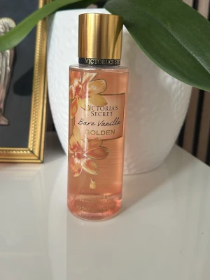 Victoria's Secret Bare Vanilla Golden - En parfym från Victoria's Secret med namnet Bare Vanilla Golden. Flaskan är cylindrisk och transparent med en gyllene skruvkork och blommigt motiv i guld och beige. Innehållet har en ljus aprikosfärg och flaskan är tillverkad av plast.