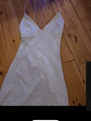 Vit ärmlös klänning med smala band - Snygg vit klänning med smala axelband och v-ringad halsringning. Klänningen är kort och har en figurnära passform, perfekt för dig som gillar en stilren och enkel look. Materialet ser ut att vara polyester eller syntet.