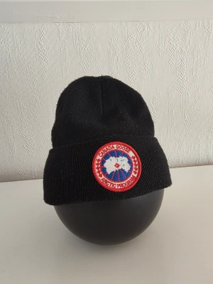 Svart mössa med patch - Svart ribbstickad mössa med uppvikt kant och rund röd-vit-blå patch framtill med logotyp och texten 'Arctic Program'. Klassisk och enkel design som håller dig varm under kalla dagar.