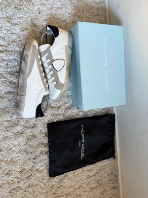 Philippe Model vita och beige sneakers - Snygga sneakers från Philippe Model i vitt och beige läder med mörkblå detaljer på hälen. Klassisk låg modell med vita skosnören och gummisula. Kommer med originalkartong och dustbag. Perfekta för en clean och trendig look.