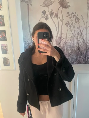 Kort svart dubbelknäppt kappa - Snygg kort svart kappa med dubbelknäppning och stora knappar. Jackan har en klassisk krage och axelklaffar som ger en trendig look. Perfekt för dig som vill ha en stilren och enkel kappa till hösten eller vintern. Andvänd en gång säljer pga för liten