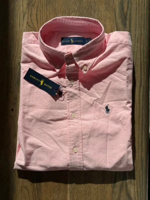 Rosa skjorta från Ralph Lauren - Klassisk rosa skjorta från Ralph Lauren med button-down krage och broderad logga på bröstet. Skjortan har långa ärmar, vita knappar och är tillverkad i mjuk bomull. Perfekt för en clean och stilren look.