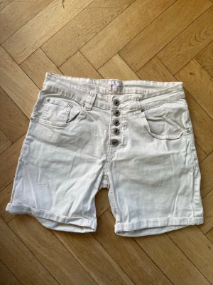 Vita denimshorts med knappar - Snygga vita shorts i denim med hög midja och fem knappar framtill. Klassiska fickor både fram och bak samt uppvikta benslut för en avslappnad look. 