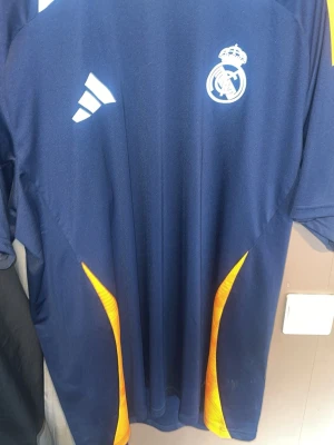 Real Madrid tränings-tshirt Adidas - Säljer en mörkblå Real Madrid tränings-tshirt från Adidas med klubbens logga på bröstet och vita Adidas-detaljer. T-shirten har orange inslag på sidorna och är tillverkad i ett lätt och funktionellt material som passar perfekt för sport och träning. 