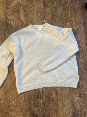 Vit stickad tröja med rund hals - Klassisk vit stickad tröja med rund halsringning och långa ärmar. Tröjan har ribbade muddar vid ärmslut och nederkant för en enkel men stilren look. Perfekt att matcha med jeans eller kjol.