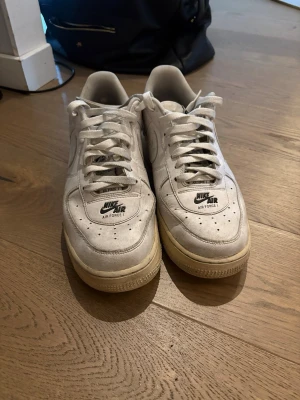 Nike Air Force 1 vita sneakers - Klassiska vita Nike Air Force 1 sneakers med svart logga på plösen och hälen. Skorna har en låg siluett, perforerad tåbox och robust gummisula. Tillverkade i läder med textildetaljer och snörning framtill. Perfekta för en streetwear-look.