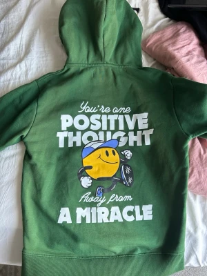Grön hoodie Seek Discomfort - Grön hoodie från Seek Discomfort med stor vit text och en glad smiley-figur på ryggen. Trycket lyder 'You're one positive thought away from a miracle'. På framsidan finns ett mindre tryck med texten 'Seek Discomfort'. Hoodien har huva och ribbade muddar.
