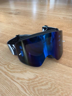 Blå spegelglas crossglasögon Raven - Säljer ett par Raven crossglasögon med blått spegelglas och bred svartvit rem med logga. Glasögonen har en böjd form och mjuk skumvaddering på insidan för extra komfort. Perfekta för motorsport och skyddar mot damm och vind.