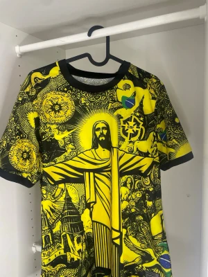 Gul och svart Brasilien t-shirt - Unik gul och svart t-shirt med stort tryck av Kristusstatyn och brasilianska symboler. T-shirten har korta ärmar, rund hals och detaljerat grafiskt mönster över hela plagget. Perfekt för dig som vill sticka ut och visa kärlek till Brasilien.