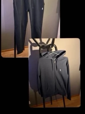 Mörkblå set från Polo Ralph Lauren - Mörkblått set med hoodie och mjukisbyxor från Polo Ralph Lauren. Hoodien har dragkedja, fickor och vit broderad logga på bröstet. Byxorna har dragsko i midjan och vit logga på benet. Båda delar är gjorda i mjuk bomull och har en clean, avslappnad stil.