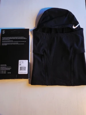 Nike Pro Therma-FIT svart balaclava - Nike Pro Therma-FIT balaclava i svart, tillverkad i ett stretchigt och värmande material. Huva med vit Nike-logga framtill, designad för att täcka huvud och hals. Perfekt för kalla dagar och sportaktiviteter utomhus.