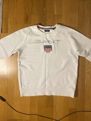 Vit sweatshirt från GANT - Säljer en vit sweatshirt från GANT med broderad logga och ett sköldmärke med stjärnor och ränder på bröstet. Tröjan har rund halsringning, långa ärmar och ribbade muddar. Perfekt för en clean och sportig look.
