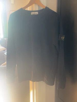 Svart långärmad tröja från Stone Island - Cool svart långärmad tröja från Stone Island med klassisk rund hals och ikonisk logopatch på vänster ärm. Tröjan är enkel och stilren, perfekt för dig som gillar streetwear och vill ha något som sticker ut lite. Materialet är mjukt och bekvämt.