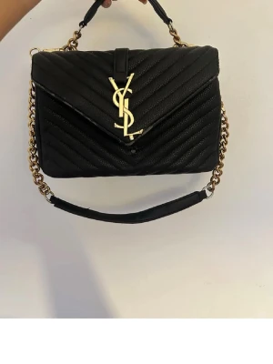 Svart YSL handväska med kedja - Stilren svart handväska från Saint Laurent med quiltad design och guldiga kedjedetaljer. Väskan har YSL-logga i metall på framsidan och ett praktiskt handtag samt axelrem. Materialet är läder och formen är rektangulär med kuvertlock.