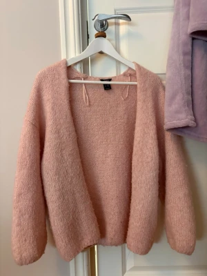 Ljusrosa stickad kofta från H&M - Mysig ljusrosa stickad kofta från Lindex med öppen front och långa ärmar. Koftan är gjord i ett fluffigt material som känns riktigt skönt och har en avslappnad passform. Perfekt att slänga på sig när det är lite kyligt.