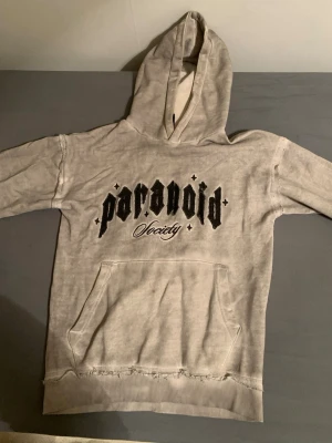 Grå hoodie Paranoid Society - Grå hoodie från Paranoid Society med svart broderad text på bröstet och stor magficka. Hoodien har en klassisk huva och ribbade muddar. Materialet ser ut att vara mjukt bomullstyg med en tvättad look. Perfekt för en avslappnad streetwear-stil.