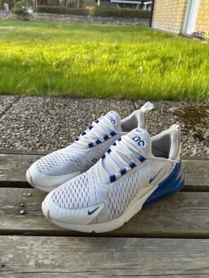 Nike Air Max 270 vita/blå sneakers - Nike Air Max 270 sneakers i vitt meshmaterial med blå detaljer och stor synlig Air-enhet i hälen. Skorna har en sportig siluett, ventilerande ovandel och klassisk Nike-logga på sidan och tungan. Perfekta för dig som gillar streetwear och bekväma sneakers.