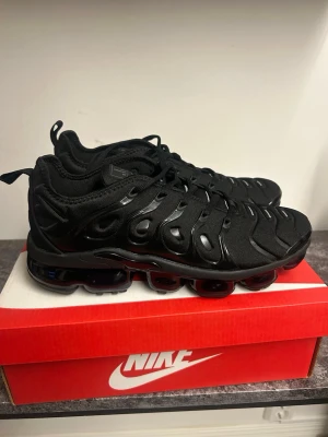 Nike Air Vapormax Plus svarta sneakers - Nike Air Vapormax Plus sneakers i helsvart med en chunky sula och ikoniska plastdetaljer längs sidorna. Ovandelen är i mesh och syntetmaterial, och skon har en sportig, futuristisk vibe. Perfekta för dig som gillar streetwear och vill sticka ut.