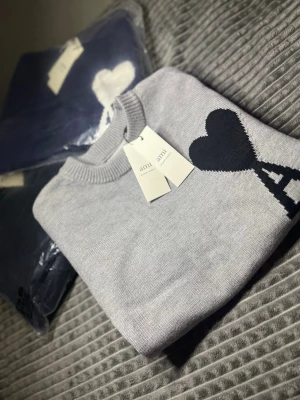 Grå stickad tröja från Ami Paris - Snygg grå stickad tröja från Ami Paris med ikonisk svart hjärta och A-logo på bröstet. Tröjan har rund halsringning och ribbade muddar vid ärmslut och nederkant. Perfekt för dig som vill ha en trendig och stilren look inför sommarkvällarna☀️🕶️