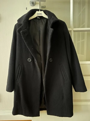 Svart ullrock från Zara - Stilren svart rock från Zara kids med dubbelknäppning och bred krage. Rocken är tillverkad i ett strukturerat ullmaterial och har två snedställda fickor framtill. Perfekt för kalla dagar och ger en klassisk look.