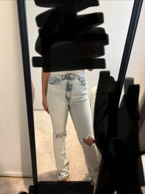 Ljusblå ripped jeans med hög midja - Säljer ett par ljusblå jeans med hög midja och raka ben. Jeansen har slitningar och hål på knäna för en trendig look. Klassisk femficksmodell och knappgylf. Perfekta för dig som gillar en avslappnad och cool stil.