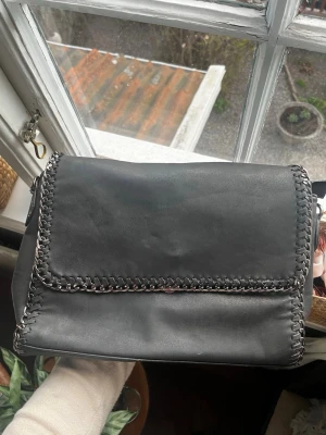 Svart axelväska med kedjedetaljer - Cool stor grå/svart handväska utan axelband. Den har snygga kedjedetaljer längs kanterna. Väskan har ett stort fack och flera innerfickor med dragkedja, perfekt för att organisera dina grejer. Materialet är slätt och ger ett stilrent intryck. Köpt på karlaplansloppisen därav vet jag inte vart den är ifrån.💞