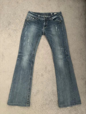 Blå bootcut jeans med broderade fickor - Snygga blå bootcut jeans från Miss me med slitningar framtill och broderade bakfickor med små diamanter, en diamant på bakfickan är borta men syns inte så mycket. Jeansen är låg midjade.