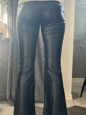 Svarta utsvängda skinnbyxor - Snygga svarta skinnbyxor med utsvängda ben och låg midja. Byxorna har en stilren look utan synliga detaljer framtill och passar perfekt för dig som gillar trendiga och tidlösa plagg. Materialet är glansigt skinn.