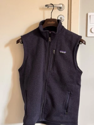 fleeceväst från Patagonia - Mörkblå fleeceväst från Patagonia med hög krage och dragkedja framtill. Västen har en bröstficka med dragkedja och två sidofickor. Perfekt att slänga på sig över en hoodie eller tröja för extra värme. Klassisk och stilren design.