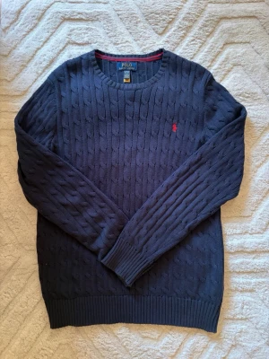 Mörkblå kabelstickad tröja Polo - Mörkblå kabelstickad tröja från Polo Ralph Lauren med rund hals och röd broderad logotyp på bröstet. Tröjan har ribbade muddar och är tillverkad i mjukt material som är perfekt för lager-på-lager. Klassisk och stilren design.