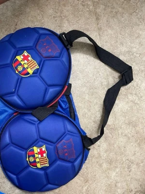 Blå FC Barcelona ryggsäck - Säljer en blå ryggsäck formad som två fotbollar med FC Barcelona-logga på båda sidor. Väskan har svarta justerbara axelband och är tillverkad i ett tåligt syntetmaterial. Perfekt för dig som älskar fotboll och vill sticka ut med en unik väska.