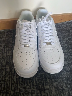 Nike Air Force 1 vita sneakers - Säljer ett par klassiska Nike Air Force 1 sneakers i helvitt läder med perforerad tå och vita skosnören. Ikonisk Swoosh på sidan och metallbricka med 'AF1' på snörningen. Perfekt för dig som vill ha en clean och tidlös look.