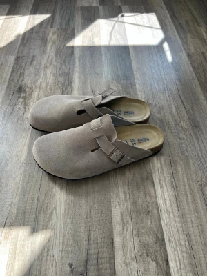 Beige Birkenstock Boston tofflor - Säljer ett par klassiska Birkenstock Boston tofflor i beige mocka med justerbart spänne och ergonomisk korksula. Perfekt för dig som vill ha både stil och komfort. Knappt använda, säljes på grund av för små för mig. Storlek 46. Priset är förhandlingsbart.