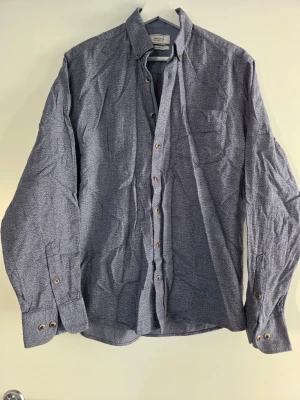 Blå skjorta från Jack's Sportswear - Snygg blå skjorta från Jack's Sportswear med comfort fit. Skjortan har diskret mönster, bröstficka och klassisk krage. Långa ärmar med knappar vid manschetterna. Perfekt till jeans eller chinos.
