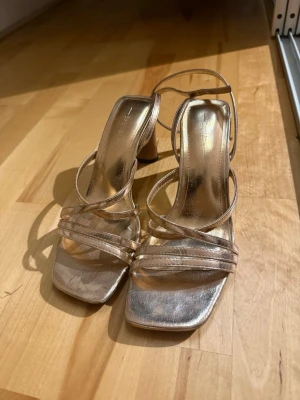 Guldiga sandaletter med klack - Säljer ett par trendiga sandaletter i metallic guld med smala remmar och fyrkantig tå. Klackarna är stabila och ger extra höjd. Perfekta för dig som vill sticka ut lite extra. Materialet är syntet med glansig finish.