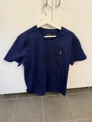 Marinblå t-shirt från Polo Ralph Lauren - Klassisk marinblå t-shirt från Polo Ralph Lauren med rund halsringning och den ikoniska broderade loggan på bröstet. Tillverkad i mjuk bomull och har en enkel, stilren design som passar till det mesta.