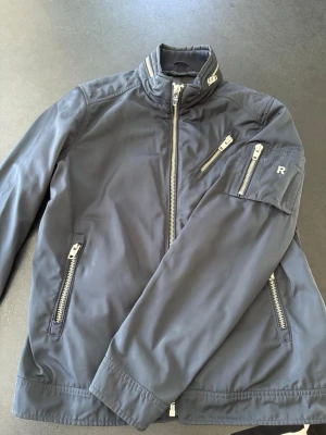 Mörkblå vindjacka Rockandblue storlek S - Snygg mörkblå vindjacka från Rockandblue med hög krage, flera dragkedjefickor och detaljer i silver. Jackan har en sportig look och är tillverkad i ett lätt och slitstarkt material. Perfekt för blåsiga dagar och streetwear-stil. Fläckfri, inga skavanker. 