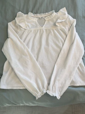 Vit blus med volangdetaljer från Kappahl - Säljer en vit blus från Kappahl med långärmad design och volangdetaljer vid axlar och halsringning. Blusen har en lätt rynkad struktur och elastiska ärmslut. Perfekt för dig som gillar en romantisk och trendig look.