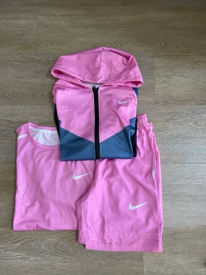 Nike rosa träningsset med hoodie - Säljer ett sportigt träningsset från Nike i rosa, bestående av en t-shirt, shorts och en hoodie med dragkedja. Alla delar har vita Nike-loggor och är gjorda i lätt, bekvämt material som passar perfekt för träning. Hoodien har även grå detaljer framtill.