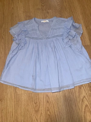Ljusblå blus med volangärmar - Säljer ljusblå blus med v-ringning och volangdetaljer på ärmarna. Blusen har korta ärmar och dekorativa sömmar upptill. Materialet är tunt och luftigt. Storlekslappen är bortklippt men är från Gina Young storlek 146/152