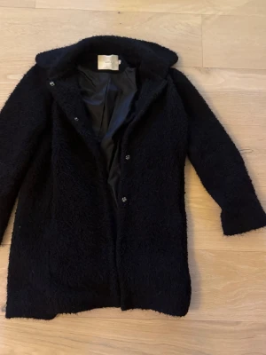 Svart kappa från Mayla - Svart kappa från Mayla med mjukt och fluffigt material. Kappan har en klassisk rak passform, knappar framtill och helsvart innerfoder. Perfekt för att hålla sig varm och snygg under kyliga dagar.