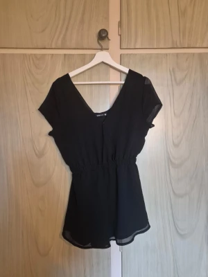 Svart playsuite - Söt svart playsuite i svalt material. Knapp så man kan justera v-rigningen om så önskas.     Alla kläder rollas innan dom skickas hem då det finns katt i hemmet🐈 Jag stryker och tvättar INTE innan jag skickar det får du stå för själv så det blir som du vill ha det🤗