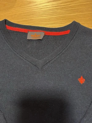 Mörkblå stickad tröja från Morris - Snygg mörkblå stickad tröja från Morris med V-ringning och diskret röd logotyp broderad på bröstet. Tröjan har långa ärmar och en röd detalj på insidan av kragen. Perfekt för dig som gillar klassisk och stilren design. Storlek S-M.