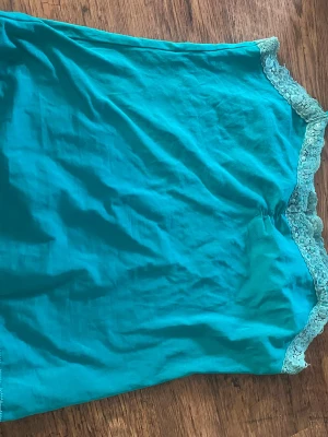 Turkost nattlinne med spets från Via Cortisa - Turkost nattlinne från Via Cortisa med tunna axelband och dekorativ spets längs halsringningen. Materialet är mjukt och lätt, perfekt för att chilla hemma. Spetsdetaljerna ger en snygg touch och nattlinnet har en enkel, rak form.