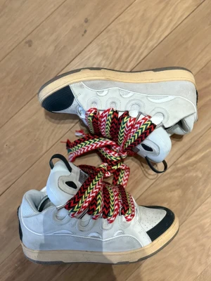 Lanvin Curb Sneakers - Unika vita curb sneakers från Lanvin med kontrasterande svart tå och häl, samt beige sula. Skorna har extra breda, färgglada skosnören i rött, grönt, svart och vitt. Ovandelen är i läder med detaljerade sömmar och perforeringar. Perfekta för dig som vill sticka ut. Bra skick och sitter skönt. Skorna är i storlek 43. Pris kan diskuteras