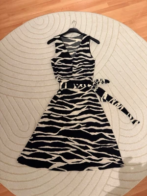Zebramönstrad omlottklänning med bälte - Snygg zebramönstrad omlottklänning i svart och vitt med v-ringning och tillhörande knytbälte i midjan. Klänningen är ärmlös och har en utställd kjol som ger en feminin siluett. Perfekt för dig som vill sticka ut med ett djurmönster.