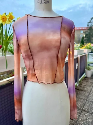 Långärmad mesh topp i pastell - Långärmad mesh topp med vågiga kanter och synliga sömmar. Färgerna går i mjuka toner av lila, orange och beige med en transparent look. Figurnära passform och supersnygg till sommaren. Över bikinin?