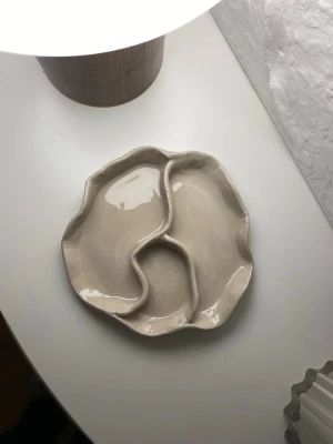 Beige skål med två fack - Unik beige skål med vågig kant och två separata fack. Skålen har en organisk form och blank yta, perfekt för snacks eller småsaker. Handgjord känsla och stilren design som passar i moderna hem. Tillverkad i lufttorkad lera av mig
